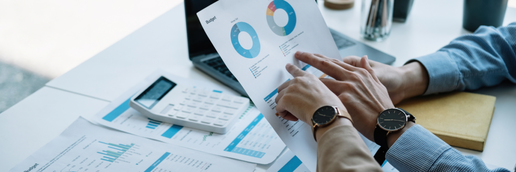 Persona analizando informe financiero con gráficos circulares y datos de marketing digital en oficina con portátil, calculadora y documentos de analítica, concepto de gestión empresarial y estrategia en España.